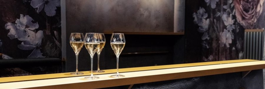 Three wine glasses stand on a bar. The background is dark with floral patterns., &copy; KESSLER Sekt GmbH & Co. KG