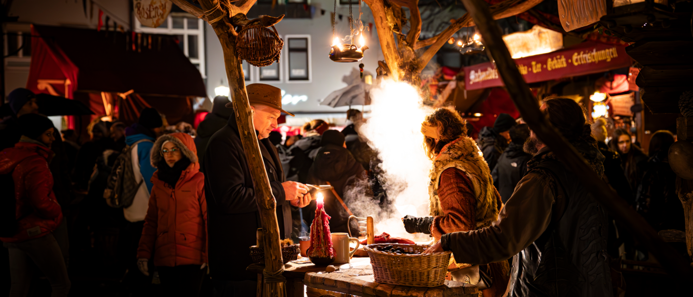 Belebter Weihnachtsmarkt mit Menschen, die an einem Stand mit Kerzen und dampfendem Essen stehen. Festliche Beleuchtung und geschäftige Atmosphäre., © Stuttgart-Marketing GmbH, Sarah Schmid