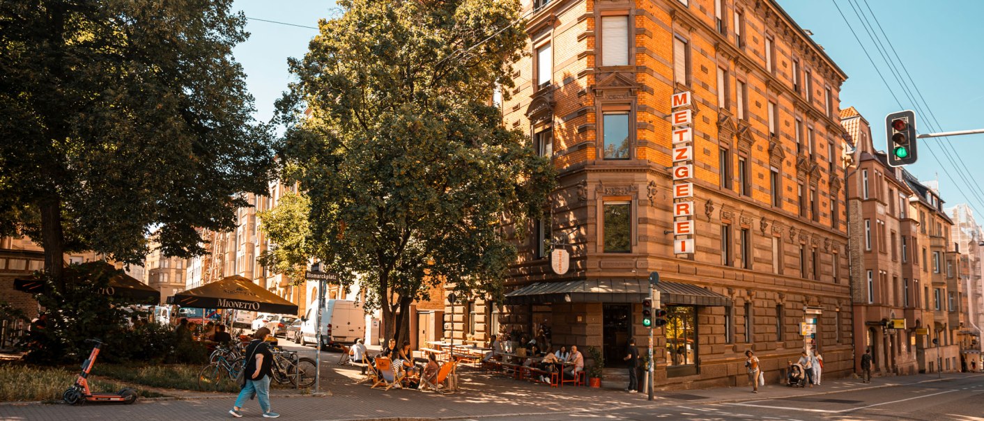 Ein belebter Platz mit einer Metzgerei, Caf&eacute;g&auml;sten unter Sonnenschirmen und einem gr&uuml;nen Ampellicht. Menschen spazieren und entspannen sich., &copy; SMG, Sarah Schmid