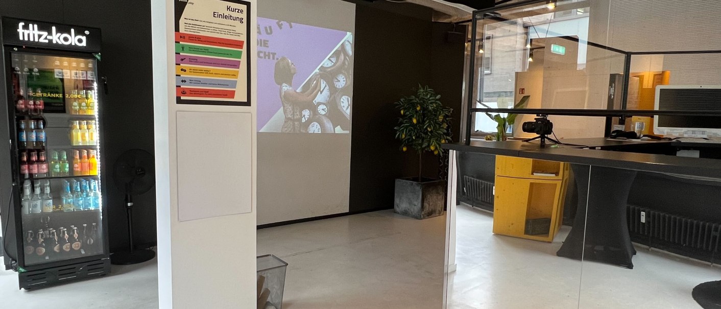 A room with a fridge, projector and reception desk. An introductory poster hangs on the wall. The room looks modern and inviting., &copy; pause & play Stuttgart