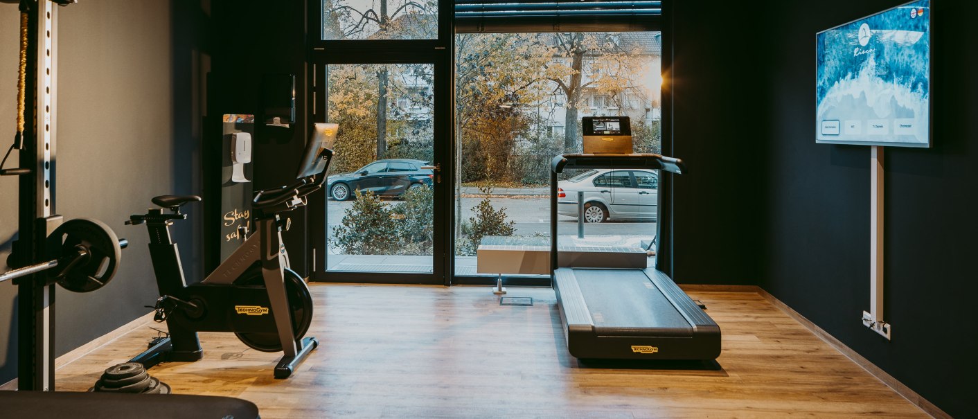 Modern fitness room with treadmill, bike and weights. Large windows offer a view of the street. A screen hangs on the wall., © Rioca Stuttgart Posto 4 GmbH Modern fitness room with treadmill, bike and weights. Large windows offer a view of the street. A screen hangs on the wall., © Rioca Stuttgart Posto 4 GmbH
