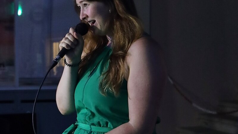 Eine Frau in einem grünen Kleid performt bei einem Poetry Slam und spricht in ein Mikrofon. Der Hintergrund ist dunkel und unscharf., © Rosenau Kultur e.V. Eine Frau in einem grünen Kleid performt bei einem Poetry Slam und spricht in ein Mikrofon. Der Hintergrund ist dunkel und unscharf., © Rosenau Kultur e.V.