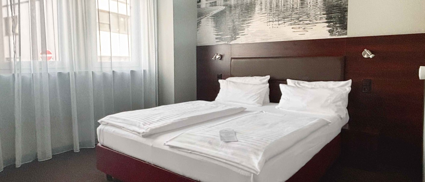 Ein gem&uuml;tliches Hotelzimmer mit einem Queensize-Bett, wei&szlig;en Bettlaken und einem gro&szlig;en Wandbild &uuml;ber dem Bett. Helle Vorh&auml;nge lassen Tageslicht herein., &copy; Novum Hospitality