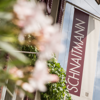 Fahne mit der Aufschrift 'SCHNAITMANN' hängt an einem Gebäude, umgeben von grünen Pflanzen und blühenden Blumen., © Weingut Schnaitmann