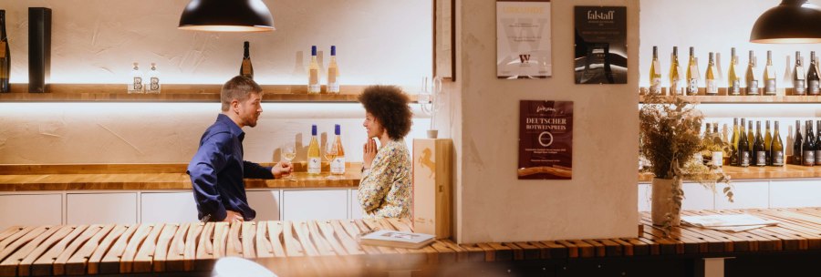 Two people are talking in a wine shop. Wine bottles on shelves, certificates on the wall. A cozy atmosphere with wooden tables., © SMG, Alwin Maigler Two people are talking in a wine shop. Wine bottles on shelves, certificates on the wall. A cozy atmosphere with wooden tables., © SMG, Alwin Maigler