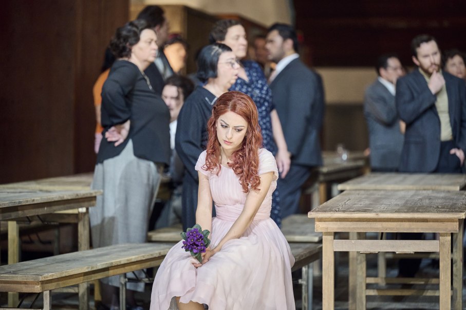 Eine Frau in einem rosa Kleid sitzt nachdenklich auf einer Bank und hält einen Blumenstrauß. Im Hintergrund stehen mehrere Personen., © Martin Sigmund