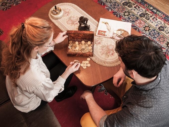 Zwei Personen sitzen an einem Tisch und betrachten eine Schachtel voller Münzen. Auf dem Tisch liegt ein Tischläufer und ein Buch., © ExitGames Stuttgart Zwei Personen sitzen an einem Tisch und betrachten eine Schachtel voller Münzen. Auf dem Tisch liegt ein Tischläufer und ein Buch., © ExitGames Stuttgart