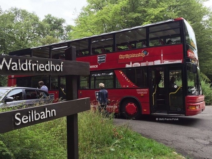 Roter Doppeldeckerbus auf einer gr&uuml;nen Stra&szlig;e, Wegweiser zum Waldfriedhof und zur Seilbahn im Vordergrund., &copy; SMG, Pierre Polak