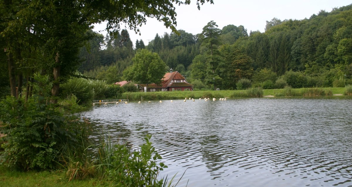 An idyllic lake with a half-timbered house in the background, surrounded by lush vegetation and trees., © Touristikgemeinschaft Hohenlohe e. V. | Susanne Schleussner An idyllic lake with a half-timbered house in the background, surrounded by lush vegetation and trees., © Touristikgemeinschaft Hohenlohe e. V. | Susanne Schleussner