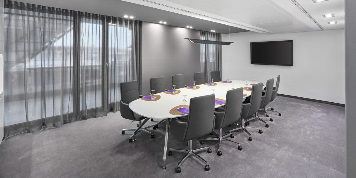 Modern conference room with oval table, gray chairs and large screen on the wall., © Jaz Hotel Stuttgart Modern conference room with oval table, gray chairs and large screen on the wall., © Jaz Hotel Stuttgart