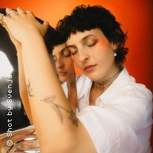 Person lehnt mit geschlossenen Augen an einem Spiegel, trägt ein weißes Hemd. Der Hintergrund ist orange. Tätowierung auf dem Arm sichtbar., © links im Bild Person lehnt mit geschlossenen Augen an einem Spiegel, trägt ein weißes Hemd. Der Hintergrund ist orange. Tätowierung auf dem Arm sichtbar., © links im Bild