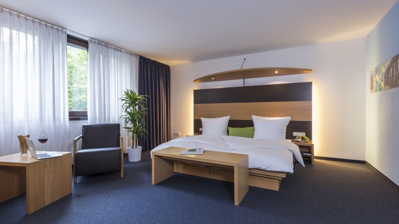 Modernes Hotelzimmer mit Doppelbett, Sessel, Tisch, Laptop und Glas Wein. Fenster mit Vorhängen, Pflanze und stilvolle Beleuchtung., © Hotel Berlin aZIS Hotel BetriebsGmbH