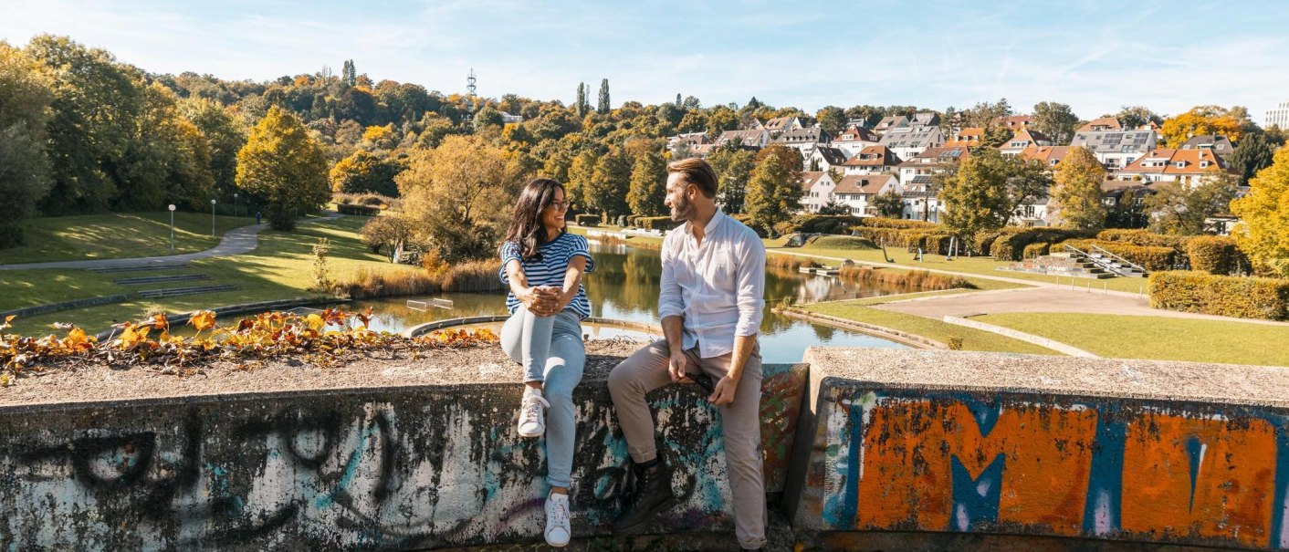 Two people are sitting on a painted wall in Wartberg Park. In the background, you can see a pond, trees, and houses. It is a sunny day., © Stuttgart-Marketing GmbH, Sarah Schmid Two people are sitting on a painted wall in Wartberg Park. In the background, you can see a pond, trees, and houses. It is a sunny day., © Stuttgart-Marketing GmbH, Sarah Schmid