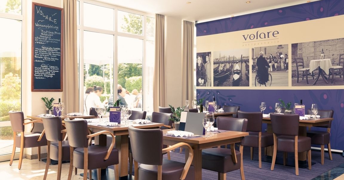 An elegant breakfast area with set tables, modern chairs, and large windows. On the wall hangs a large painting featuring the word &ldquo;Volare.&rdquo;, &copy; Mercure Hotel Stuttgart Gerlingen