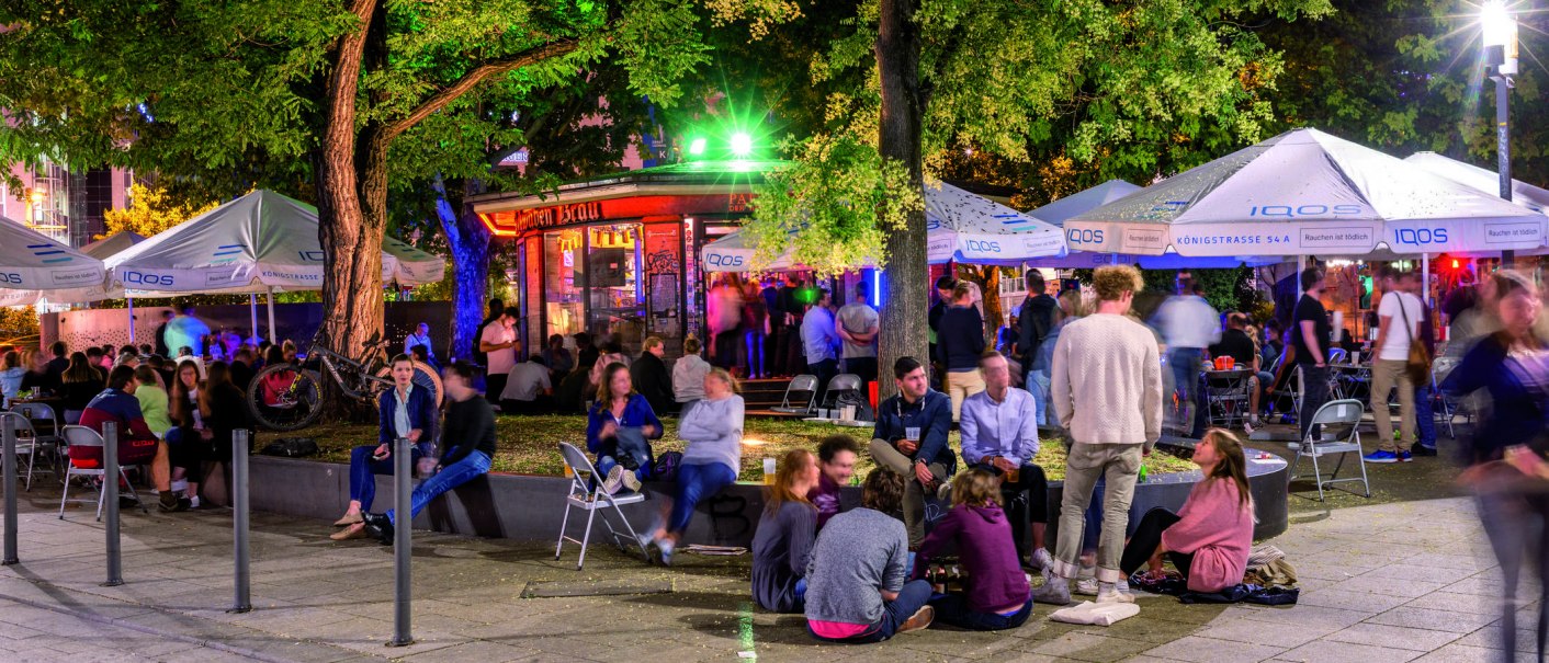 Lively outdoor area with people under illuminated umbrellas and trees. Some are sitting on chairs, others on the ground. Colorful lights in the background., © TMBW, Gregor Lengler