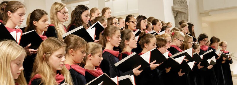 Ein Chor junger Frauen singt in einer Kirche, alle in schwarzer Kleidung mit roten Schals, halten Notenblätter., © Stiftsmusik Stuttgart