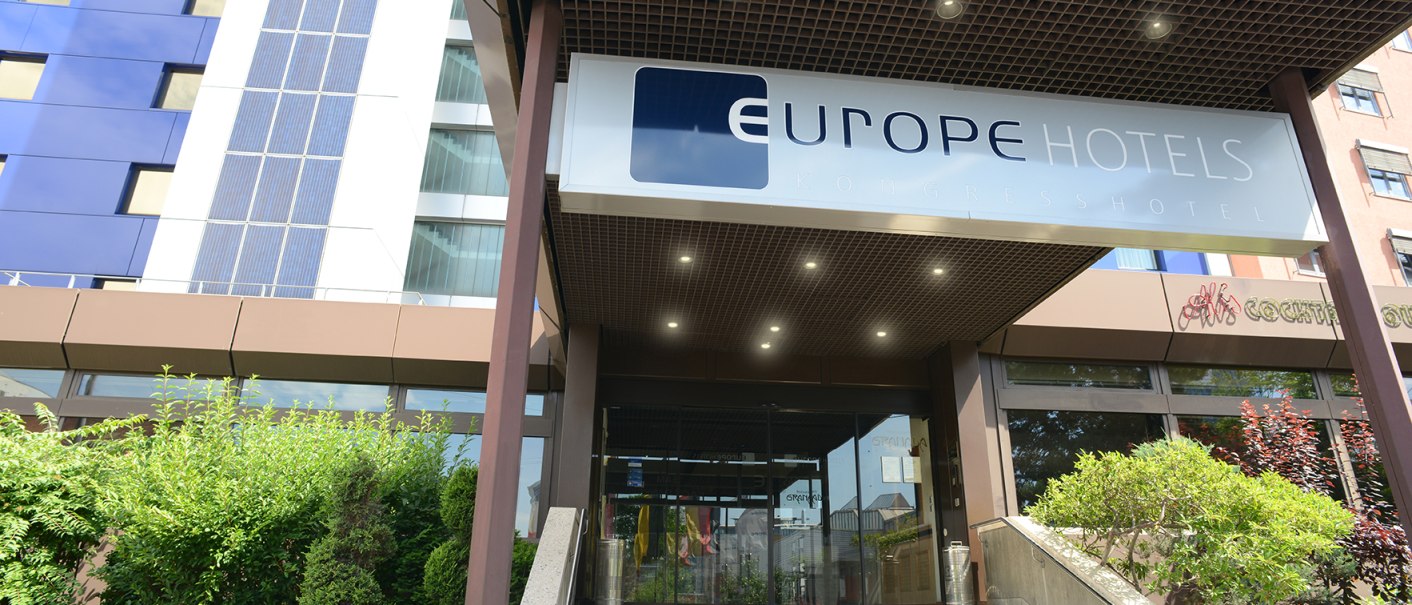 The entrance to a modern hotel with a sign reading “Europe Hotels Kongresshotel” above the stairs. Green bushes can be seen on the left., © Europe Hotels International The entrance to a modern hotel with a sign reading “Europe Hotels Kongresshotel” above the stairs. Green bushes can be seen on the left., © Europe Hotels International