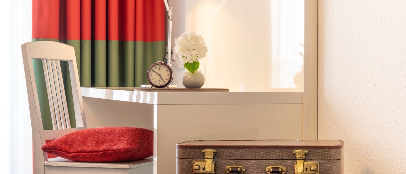 A cozy hotel room with a white desk, a red cushion on a chair, a table lamp and an old suitcase on a stool., © Hotel Weinstadt