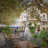 A bistro on Wilhelmsplatz with empty tables and chairs outside, surrounded by green plants and trees., © Martina Denker A bistro on Wilhelmsplatz with empty tables and chairs outside, surrounded by green plants and trees., © Martina Denker
