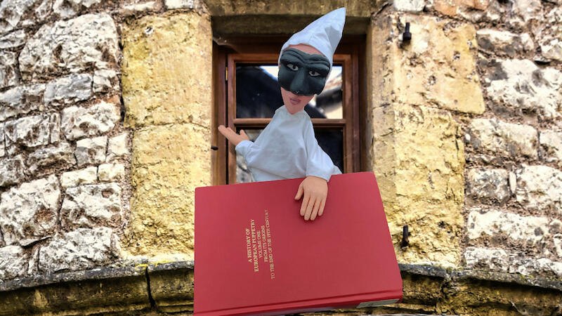 Eine Handpuppe mit schwarzer Maske und wei&szlig;em Kost&uuml;m h&auml;lt ein rotes Buch vor einem alten Steingeb&auml;ude mit Fenster., &copy; FITZ Das Theater animierter Formen