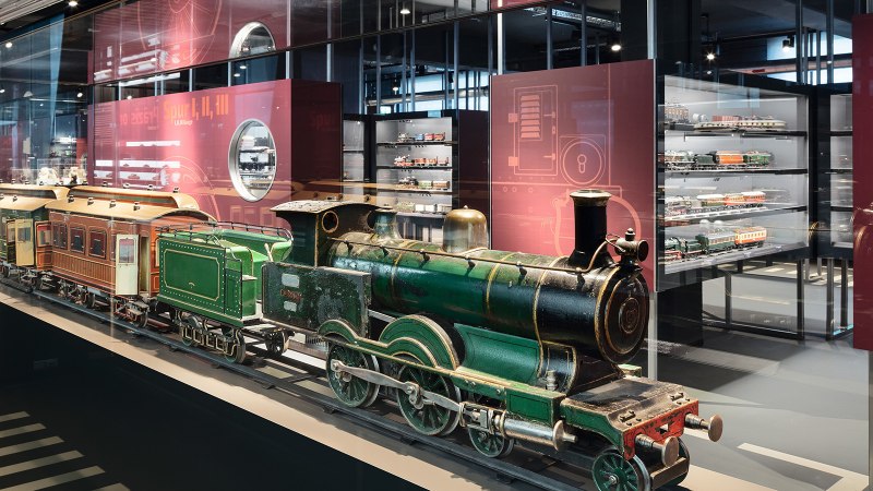 Eine grüne Modelllokomotive in einer Glasvitrine, umgeben von weiteren Modelleisenbahnen in einem Museumsausstellungsraum., © Märklineum