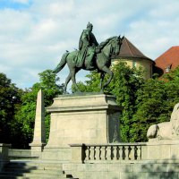 Equestrian statue on Karlsplatz in Stuttgart, flanked by lion statues and obelisks, surrounded by trees and a historic building in the background., © Stuttgart-Marketing GmbH Equestrian statue on Karlsplatz in Stuttgart, flanked by lion statues and obelisks, surrounded by trees and a historic building in the background., © Stuttgart-Marketing GmbH