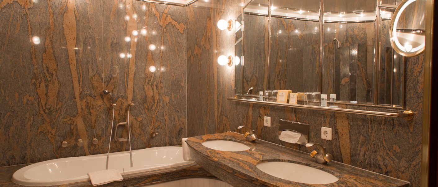 Luxuriöses Badezimmer mit Marmorwänden, Doppelwaschbecken und Badewanne. Helle Beleuchtung und Spiegel verleihen dem Raum Eleganz., © Hotel Royal