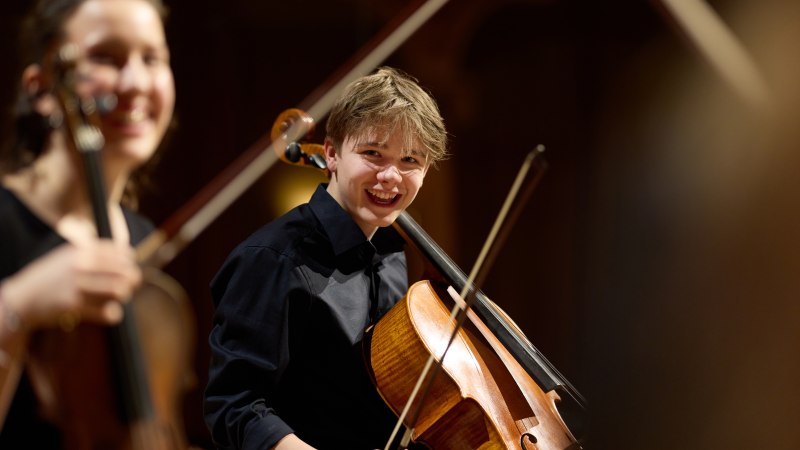 Ein junger Cellist l&auml;chelt w&auml;hrend eines Auftritts auf der B&uuml;hne. Im Hintergrund ist eine weitere Musikerin mit einer Geige zu sehen., &copy; Oliver Borchert