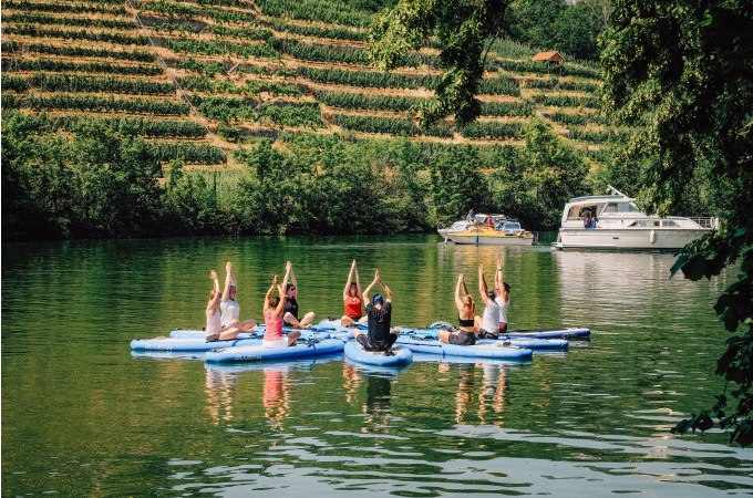 Menschen praktizieren Yoga auf SUP-Boards auf einem Fluss, umgeben von Weinbergen und einem vorbeifahrenden Boot., © Cool-Tours StattReisen Menschen praktizieren Yoga auf SUP-Boards auf einem Fluss, umgeben von Weinbergen und einem vorbeifahrenden Boot., © Cool-Tours StattReisen