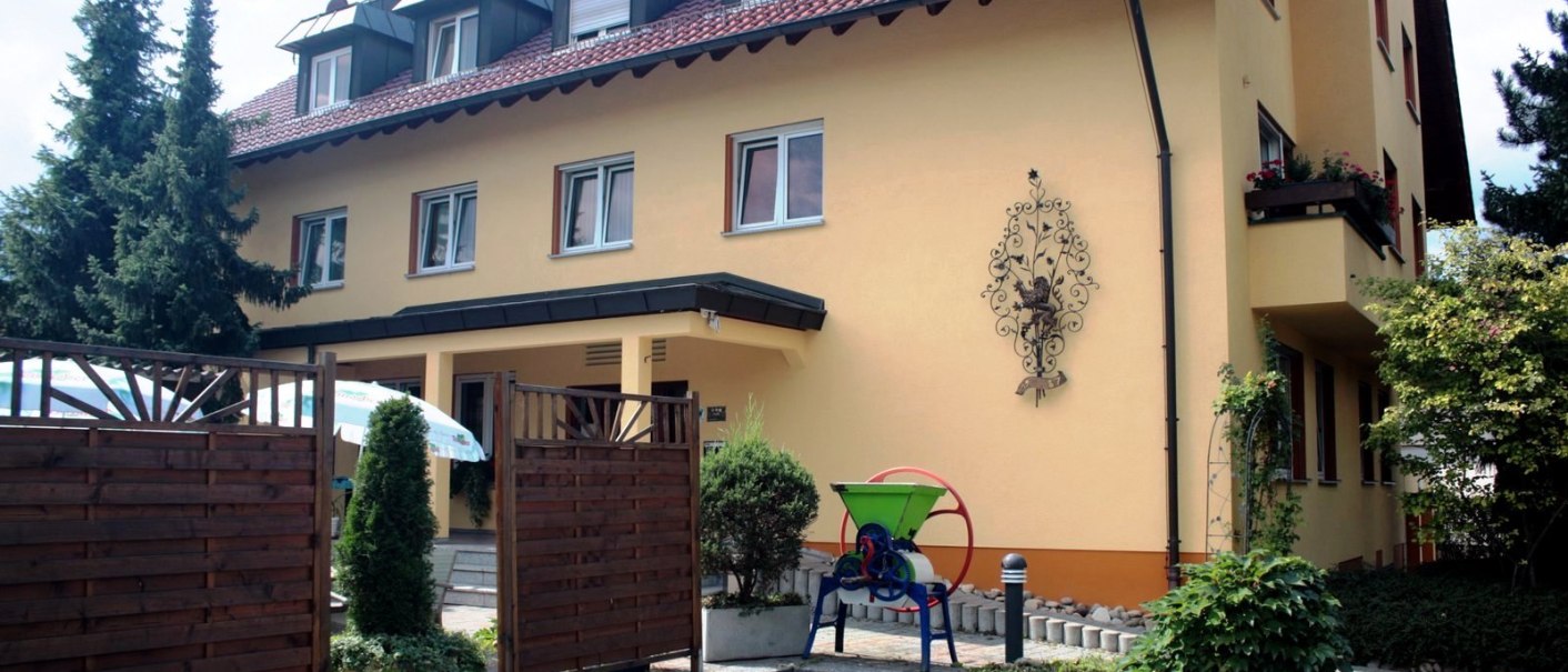 Yellow building with a red roof and hotel signage. A wooden fence and plants in front of the entrance. Decorative wall ornament and green machine in the foreground., © TOMAS Yellow building with a red roof and hotel signage. A wooden fence and plants in front of the entrance. Decorative wall ornament and green machine in the foreground., © TOMAS