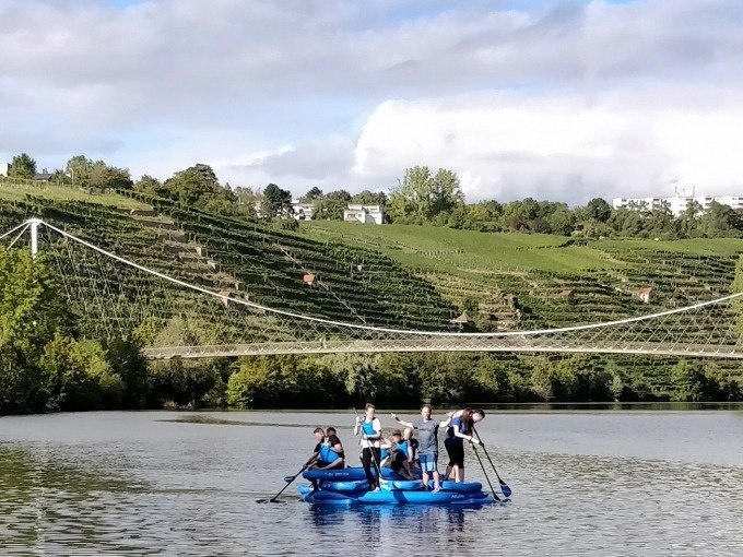 Gruppe von Menschen beim Stand-Up-Paddling auf einem Fluss. Im Hintergrund sind Weinberge und eine Hängebrücke zu sehen., © Cool-Tours StattReisen Gruppe von Menschen beim Stand-Up-Paddling auf einem Fluss. Im Hintergrund sind Weinberge und eine Hängebrücke zu sehen., © Cool-Tours StattReisen