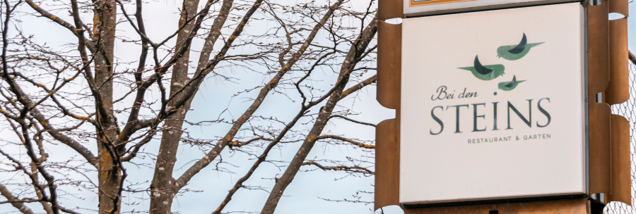 A sign for the restaurant 'Bei den Steins' with birds as a logo, in front of a bare tree and blue sky., © Stuttgart-Marketing GmbH, Sarah Schmid A sign for the restaurant 'Bei den Steins' with birds as a logo, in front of a bare tree and blue sky., © Stuttgart-Marketing GmbH, Sarah Schmid