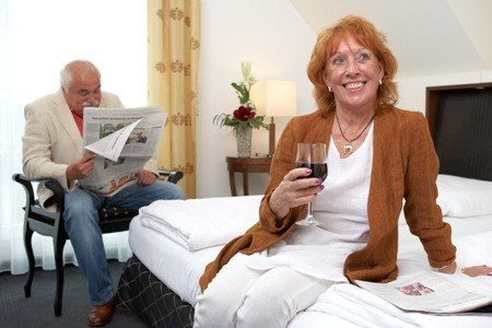 Älteres Paar in einem Hotelzimmer. Frau sitzt auf dem Bett mit Wein, Mann liest Zeitung. Gemütliche Atmosphäre., © Jeanette Bender