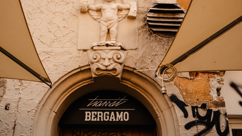 Steinrelief eines Kindes &uuml;ber einem Eingang mit der Aufschrift 'transit Bergamo'. Der Putz ist br&ouml;ckelig, Graffiti und Markisen sind sichtbar., &copy; SMG_Sarah Schmid