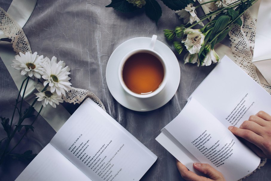 Eine Tasse Tee steht auf einem Tisch mit Blumen und einem geöffneten Buch. Eine Person blättert darin., © Foto: pexels