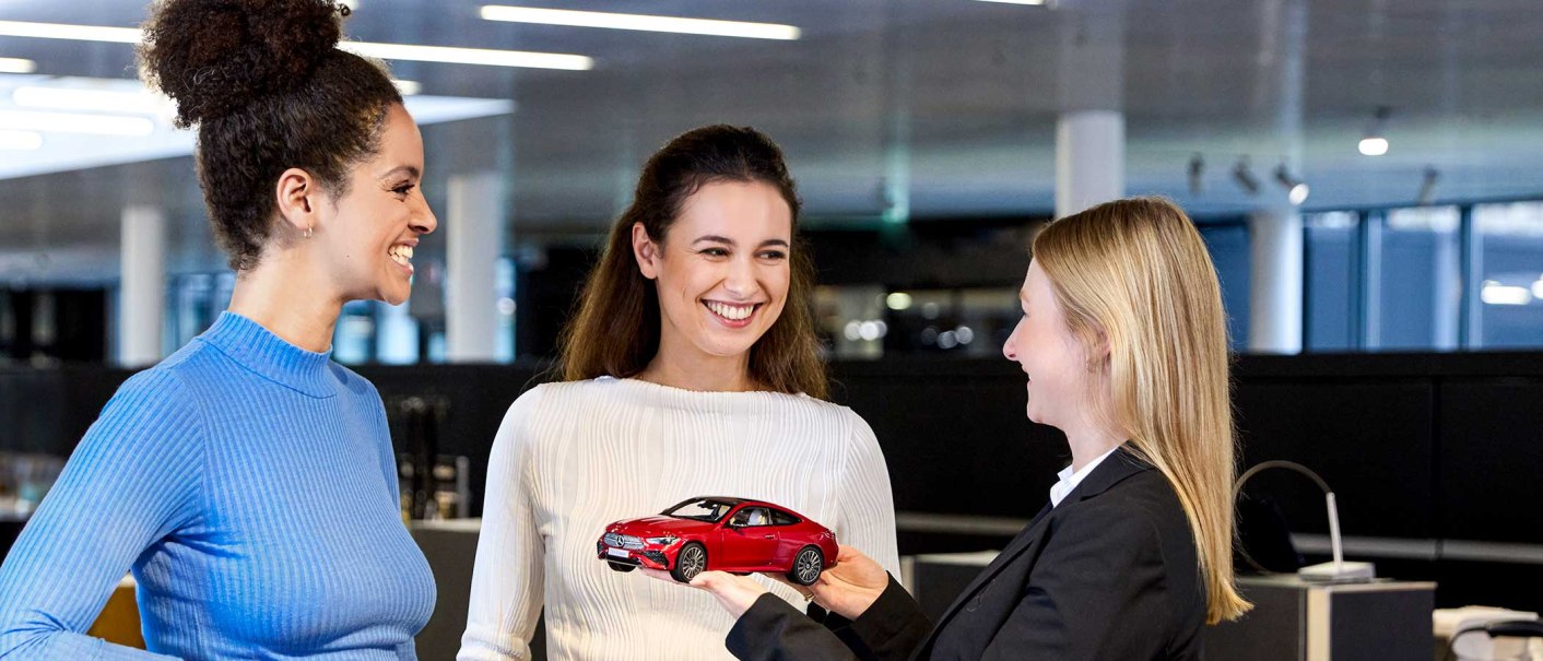 Three women smiling in a modern office. One woman is holding a red model car in her hands., © Mercedes-Benz AG