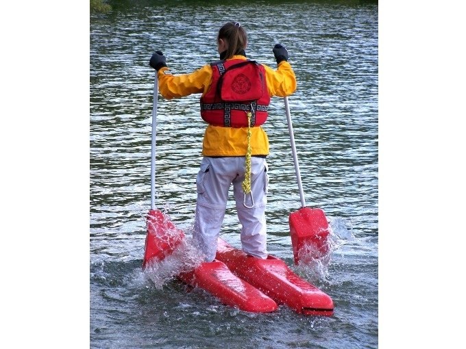 Person mit gelber Jacke und roter Schwimmweste steht auf roten Wasserschuhen im Wasser und h&auml;lt zwei St&ouml;cke., &copy; Cool-Tours StattReisen