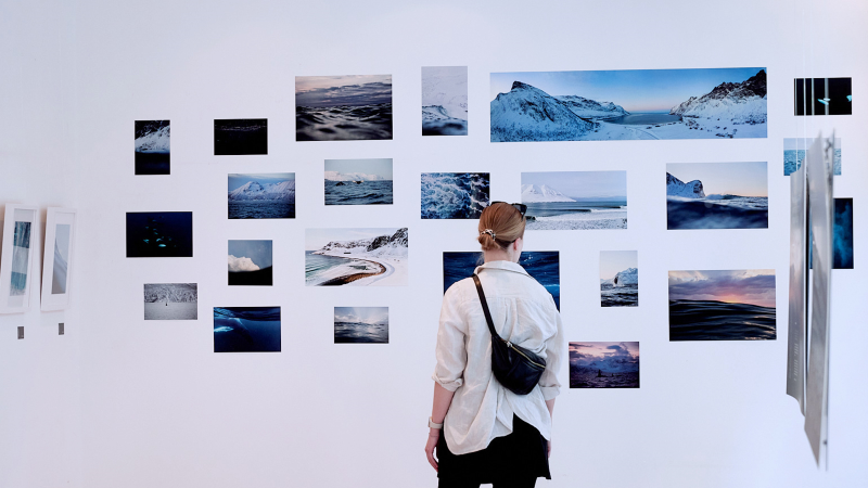 Eine Person betrachtet eine Wand mit Fotografien von Landschaften und Wasser. Die Bilder zeigen verschiedene Naturmotive in k&uuml;hlen Farbt&ouml;nen., &copy; OCEAN GALLERY