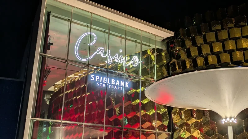 The Stuttgart Casino at night: an illuminated glass facade, a golden cube-shaped structure, and a striking white canopy., © SMG The Stuttgart Casino at night: an illuminated glass facade, a golden cube-shaped structure, and a striking white canopy., © SMG
