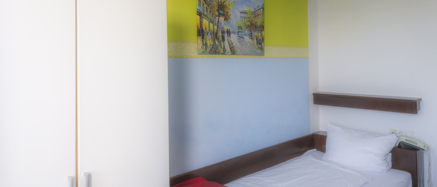 Einzelzimmer mit Einzelbett, weißem Schrank und Gemälde an der Wand. Ein rotes Tuch liegt auf dem Bett., © Hotel Ri
