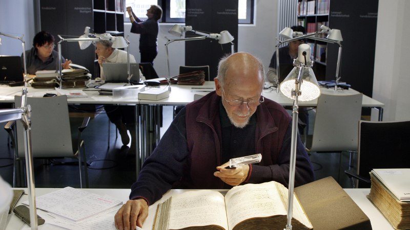 Im Lesesaal des Stadtarchivs arbeiten Menschen an Tischen mit alten B&uuml;chern und Laptops. Eine Person nutzt eine Lupe, um ein Buch zu lesen., &copy; Stadtarchiv Stuttgart; Foto: Susanne Kern