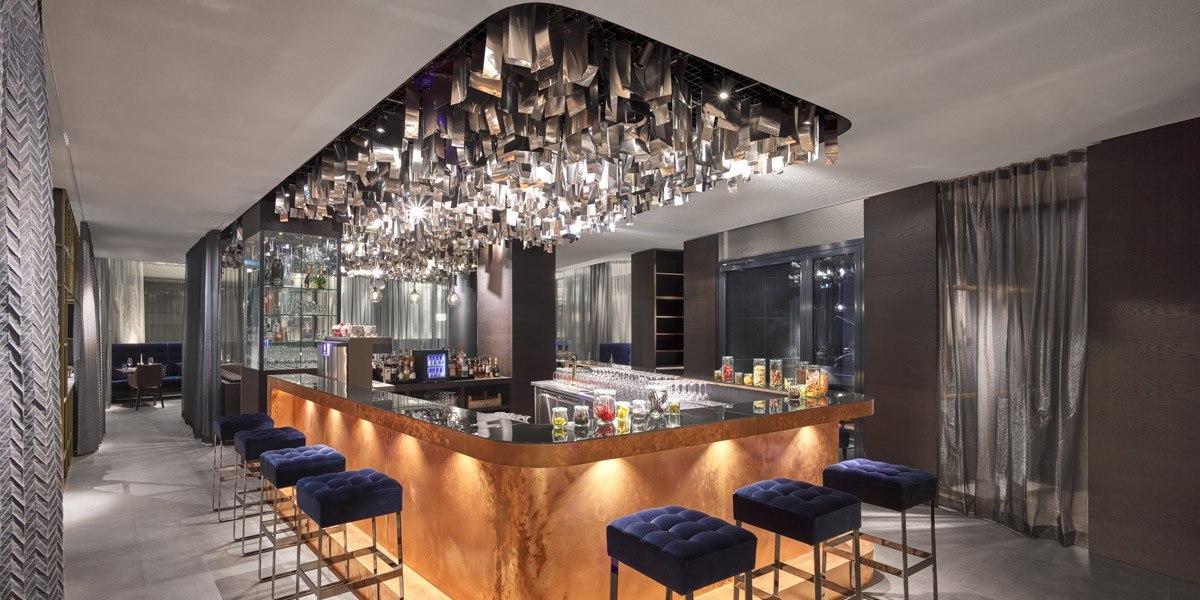 Elegant bar with illuminated counter, blue stools and an eye-catching metal ceiling installation. Stylish, modern atmosphere., © Jaz Hotel Stuttgart Elegant bar with illuminated counter, blue stools and an eye-catching metal ceiling installation. Stylish, modern atmosphere., © Jaz Hotel Stuttgart