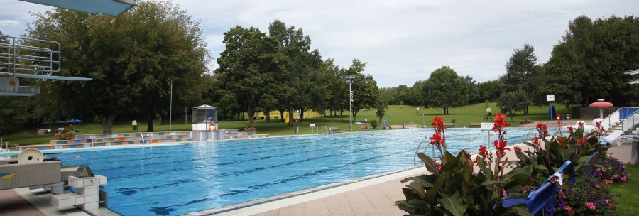 Ein Freibad mit einem großen Schwimmbecken, umgeben von grünen Bäumen und bunten Blumenbeeten. Der Himmel ist leicht bewölkt., © Stuttgarter Bäder