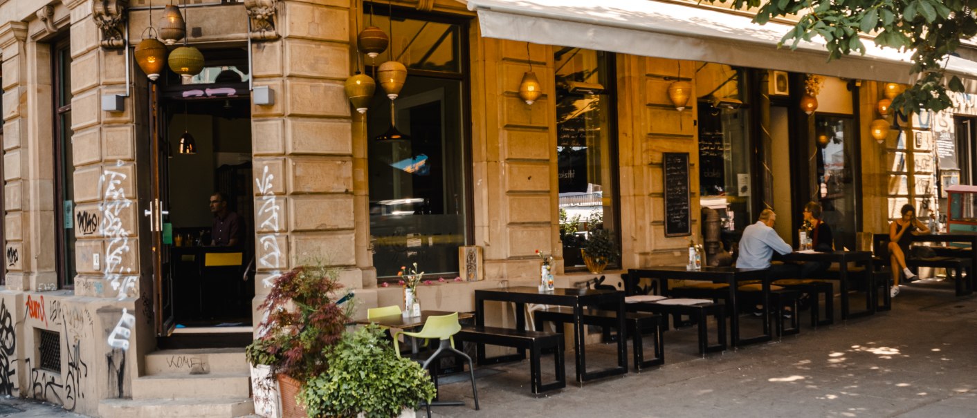Ein Café an einer Straßenecke mit Außenbestuhlung, Pflanzen und hängenden Lampen. Menschen sitzen an Tischen unter einem Sonnenschutz., © SMG, Sarah Schmid