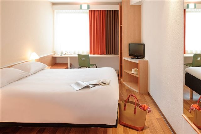 Helles Hotelzimmer mit Doppelbett, Schreibtisch, Fernseher und Tasche auf dem Boden. Vorhänge in Rot- und Brauntönen., © TOMAS Helles Hotelzimmer mit Doppelbett, Schreibtisch, Fernseher und Tasche auf dem Boden. Vorhänge in Rot- und Brauntönen., © TOMAS