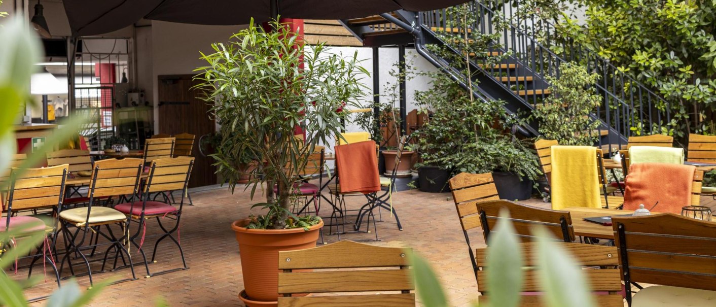 Cozy outdoor café area with wooden tables and chairs, surrounded by plants. Colorful blankets on the chairs create an inviting atmosphere., © SMG, Sarah Schmid