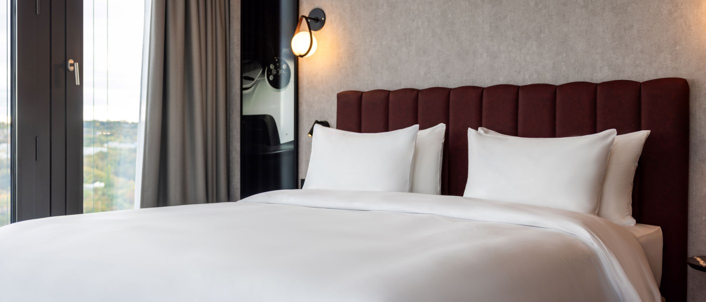 Ein elegantes Hotelzimmer mit einem gro&szlig;en Bett, wei&szlig;en Bettlaken und einem roten Kopfteil. Links befindet sich ein Fenster mit grauen Vorh&auml;ngen., &copy; Radisson Hotel Group