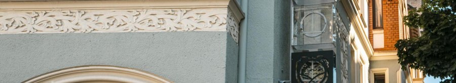 Fassade eines Gebäudes mit verzierten Elementen und einem Schild mit Logo. Ein Baum wirft Schatten auf die Wand. Klare, sonnige Atmosphäre., © SMG, Sarah Schmid