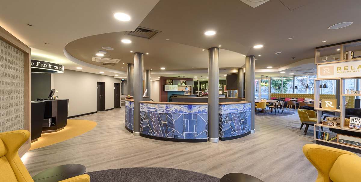 Moderne Hotellobby mit runder Rezeption, gelben Sesseln und Regalen. Helle Beleuchtung und stilvolle Einrichtung schaffen eine einladende Atmosphäre., © TOMAS