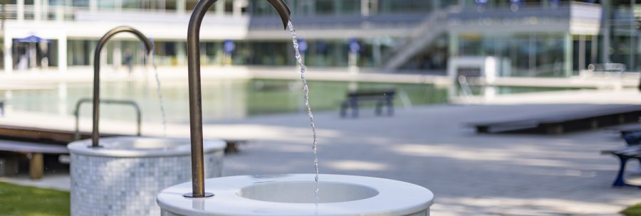 A drinking fountain in Mineralbad Berg with running water in the foreground, a modern building and a pool in the background., © Stuttgarter Bäder